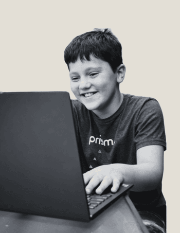 young Prisma online homeschool learner wearing a Prisma t-shirt smiling while working on a laptop