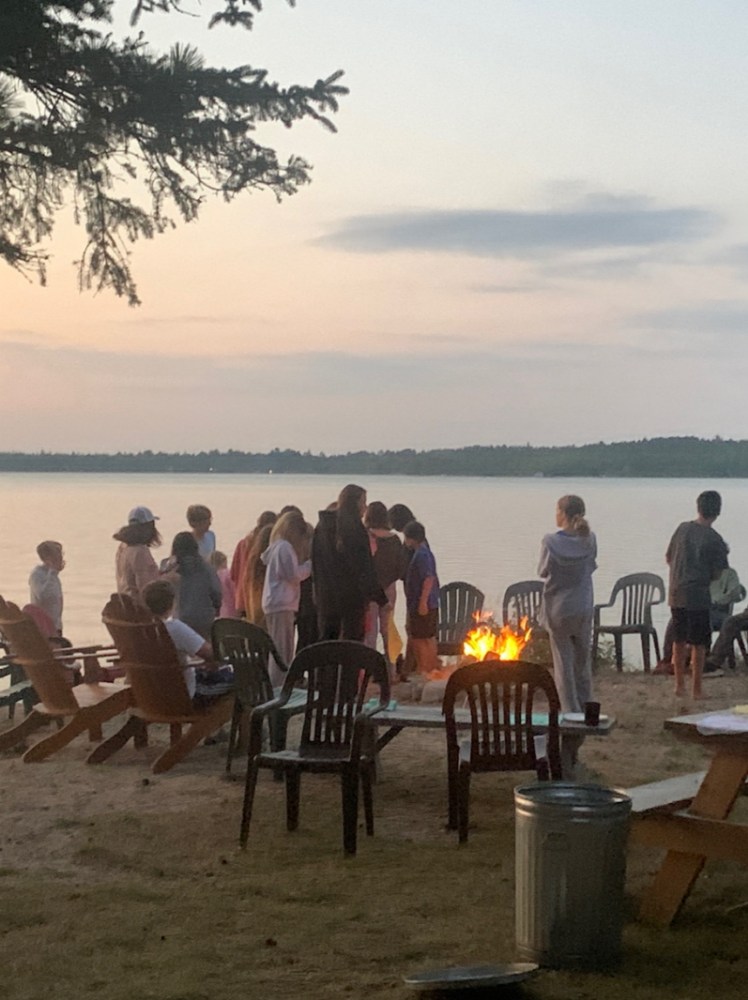 Prisma families gathered around a campfire by a lake at sunset