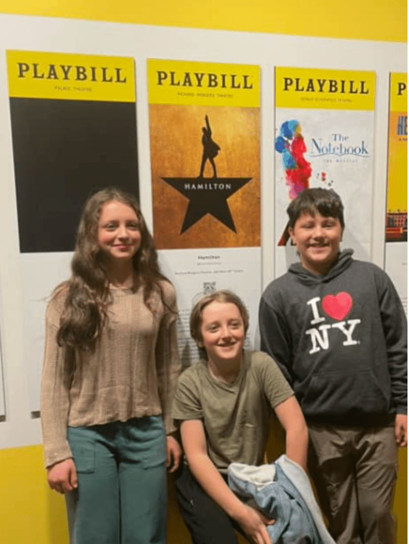 three Prisma middle school students smiling in front of Broadway Playbill posters including Hamilton