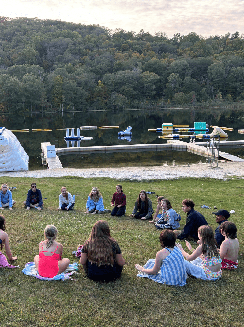 Prisma students sitting in a circle on grass by a lake during a meetup