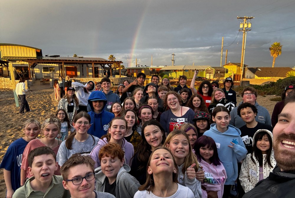 large group of Prisma students posing for a selfie on a beach with a rainbow