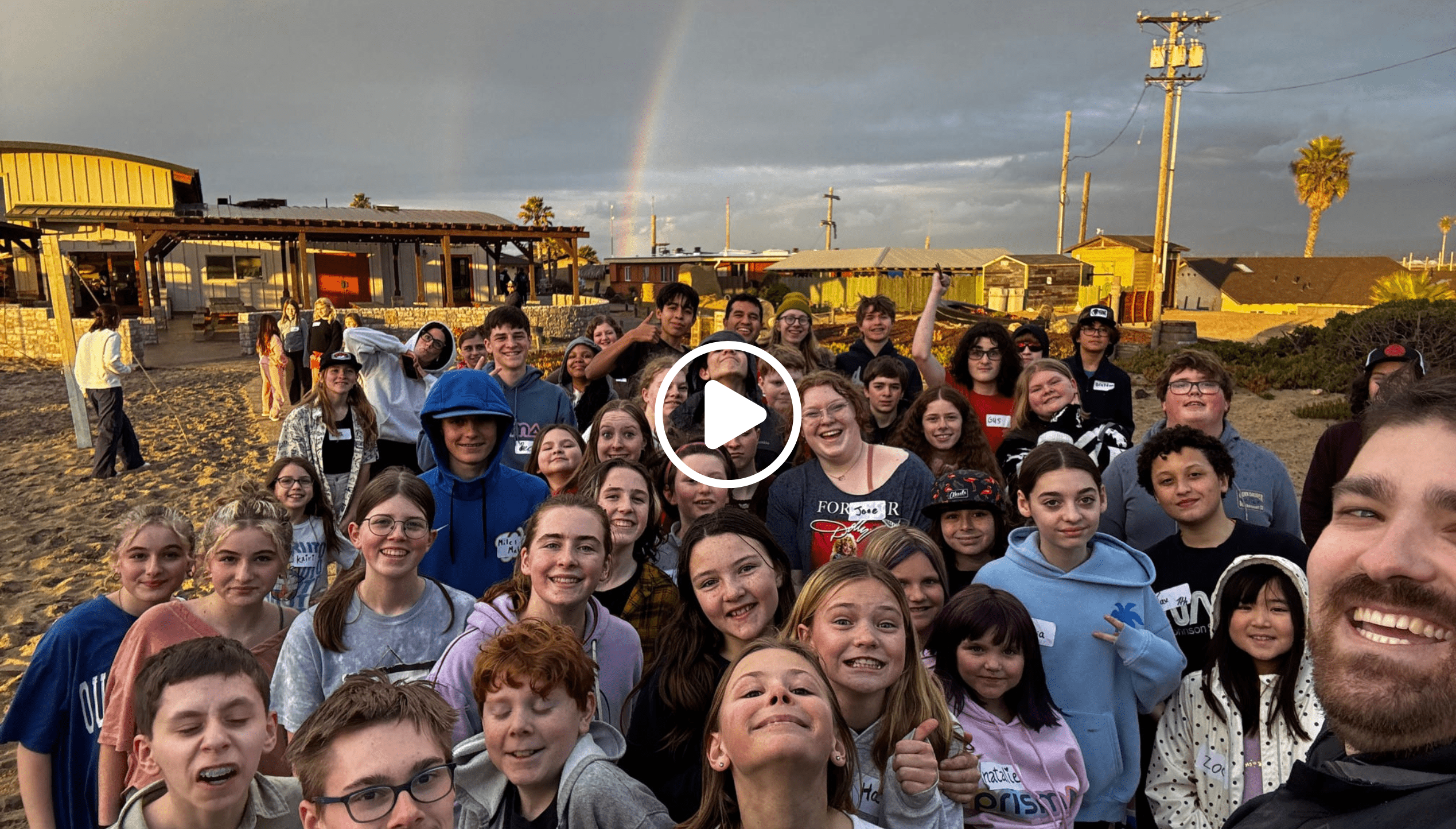 large group selfie of Prisma learners smiling outdoors with a rainbow in the background