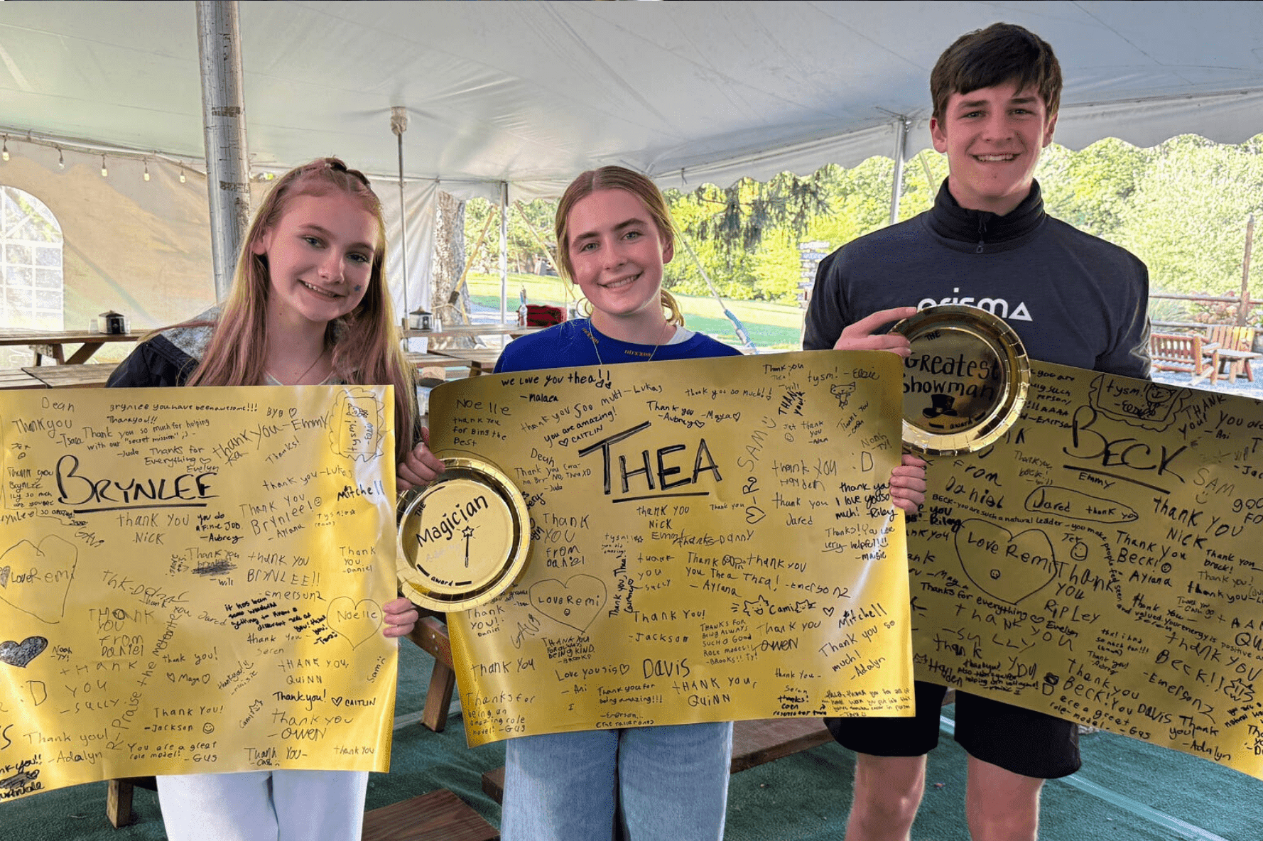 three Prisma high school students holding signed gold award posters under a tent